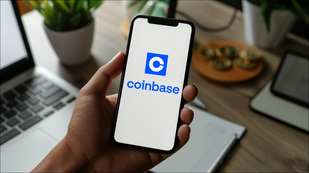 Eine Hand hält ein Smartphone mit dem Coinbase-Logo auf dem Bildschirm, umgeben von einer Büroumgebung mit Laptop und physischen Bitcoins im Hintergrund.