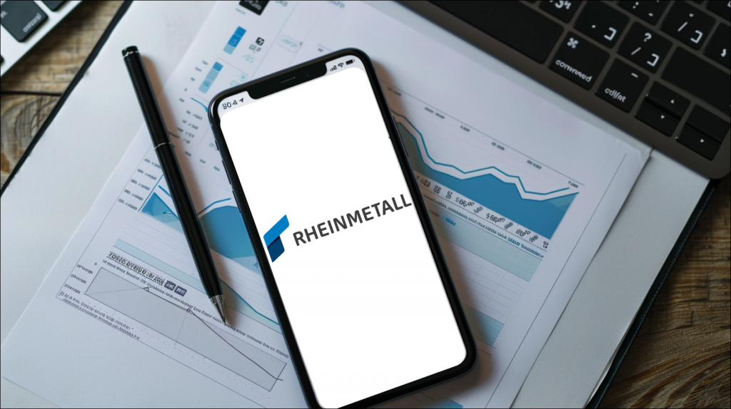 Ein Arbeitsplatz mit einem Smartphone, auf dessen Display das Logo von RHEINMETALL, neben Papieren mit Diagrammen