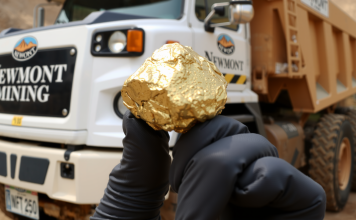 Ein glänzender Goldnugget in einer behandschuhten Hand vor einem Newmont Mining-Lastwagen